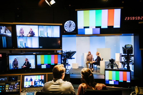 Two crew work in a TV production studio surrounded by cameras, screens and presenters